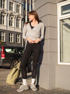 Woman leaning against a brick wall outside wearing a wrap-a-round top in grey melange color. En kvinna står lutat mot en tegelvägg utomhus klädd i en gråmelerad omlott top.